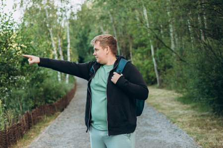 One Caucasian male hiking in the woods in summer. Travel, active lifestyle, people and hobby concept. Blonde male in sport clothes pointing finger outdoors. Traveler enjoys eco-tourism.の写真素材