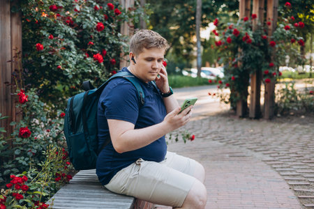 Bottom view smiling happy fun young man in blue t-shirt sit on bench use mobile cell phone chat...