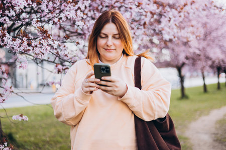 Happy 30s woman cheerfully talking on a smartphone against a background of pink tree blossoms. Spring time in the city.の写真素材