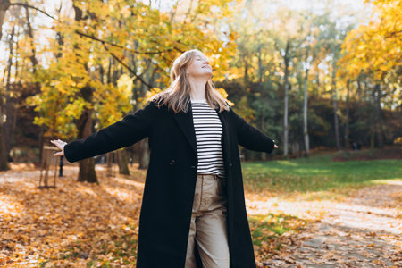 30s blonde Girl in motion expressing happiness at city park. Attractive, cheerful happy woman raising hands up, feeling free and joyful during the nice autumn day.の写真素材