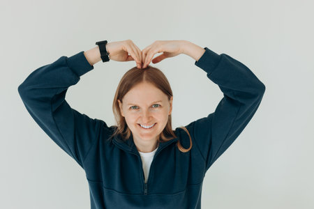 Smiling young girl in sweater showing heart with two hands, love sign. Woman standing Isolated over...