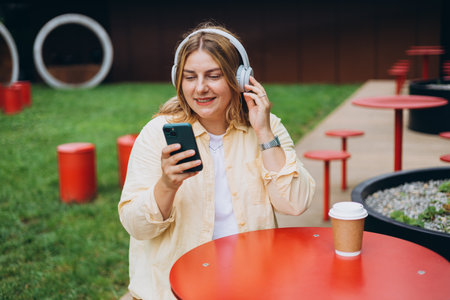 Happy young Caucasian woman in good mood listening to music with headphones and sitting on terrace street cafe in the city. Music lover enjoying music. Urban lifestyle concept.の写真素材