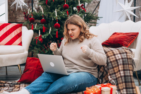 Stressed angry young woman working on laptop, entering wrong payment information, purchasing New year gifts, sitting on the floor near decorated Christmas tree.の写真素材