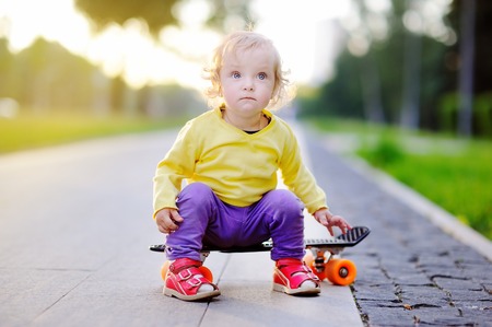 Cute toddler girl sitting on skateboard on summer sunny streetの写真素材