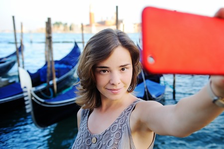 Young female traveler making selfie photo using her smartphone standing near San Marco square with gondolas on the background in Venice, Italyの写真素材