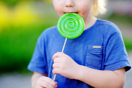 Child with allergic reaction eating big green lollipop. Unhealthy sweet food for young kids. Allergic rash dermatitis on toddler skin face.の写真素材