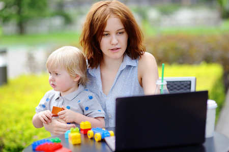 Young mother working oh her laptop and holding her sad toddler sonの写真素材