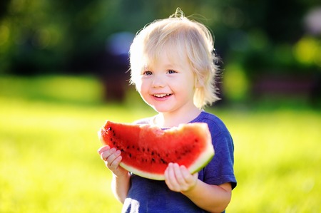 Cute caucasian little boy with blond hairs eating fresh watermelon outdoorsの写真素材