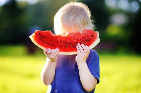Cute caucasian little boy with blond hairs eating fresh watermelon outdoorsの写真素材