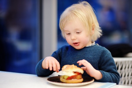 Cute blonde boy eat hamburger at fast food restaurant. Child unhealthy meal conceptの写真素材