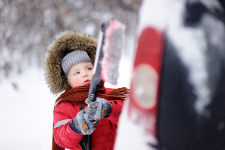 Cute little child helping to brush a snow from a car. Toddler boy using tool for cleaning fathers carの写真素材