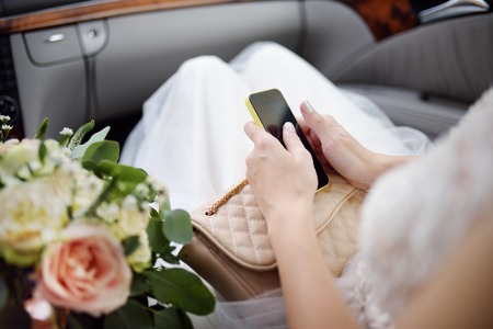 Close-up photo of bride sitting in car and holding her smartphone during her wedding day....
