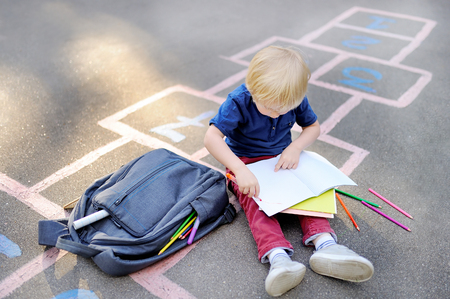 Cute blond boy doing homework sitting on school yard after school with bags laying near. Back to school concept. Little boy on school yardの写真素材