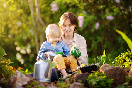 Beautiful young woman and her cute son planting seedlings in bed in the domestic garden at summer day. Garden tools, gloves and watering can outdoors. Gardening activity with little kid and familyの写真素材