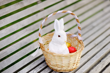 Colorful Easter eggs in a basket on wooden bench with cute white toy bunny. Celebrating Easter outdoors. Accessories for Easter party in parkの写真素材