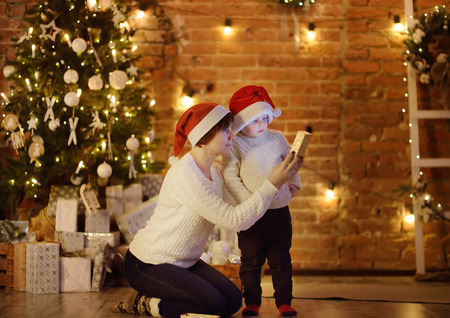 Cute little boy wearing Santa hat and his mother or grandmother regard a Christmas gift. Portrait of happy family on Christmas eve. Cozy decorated living roomの写真素材