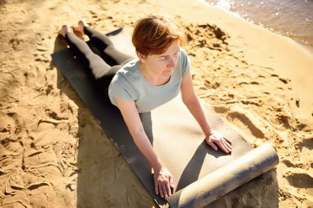 Mature woman practicing yoga outdoor exercise on the beach near the river. Healthy lifestyle.