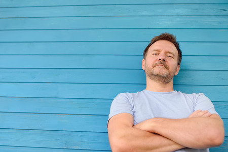 Shot of handsome mature man crossing hands and look at camera. Male portrait with blue wooden wall on background.の写真素材