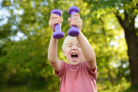 Cute little boy lifting dumbbells outdoors. Kid's sport.