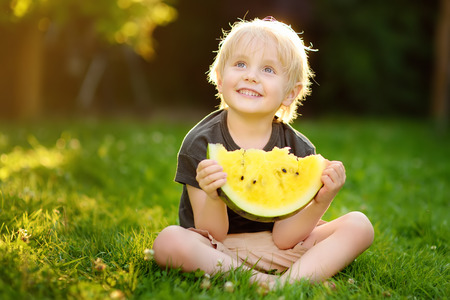 Cute caucasian little boy with blond hairs eating yellow watermelon outdoors. Seasonal fruits and vegetables for kids.の写真素材