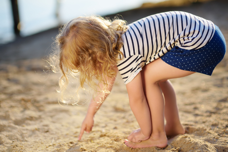 Little girl playing with sand during walk by the lake at summer day. Baby draw finger on sand. Active family time on nature.の写真素材