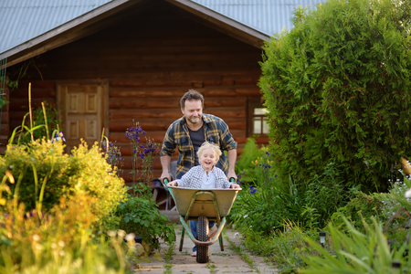 Happy little boy having fun in a wheelbarrow pushing by dad in domestic garden on warm sunny day. Active outdoors games for kids in summer.の写真素材