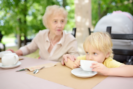 Beautiful senior lady with his little grandson drinking tea in outdoors cafe or restaurant. Elderly lady lifestyle. Grandmother and grandchild.の写真素材