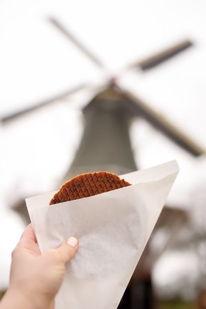Woman's hand with traditional dutch food - waffles with caramel on background of dutch windmill, Netherlandsの写真素材