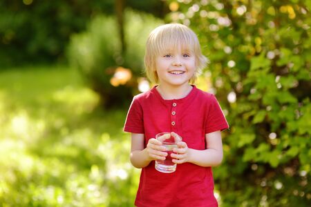 Little boy drinking glass of water in hot sunny summer day on the backyard or home garden. Healthy lifestyle for children.の写真素材