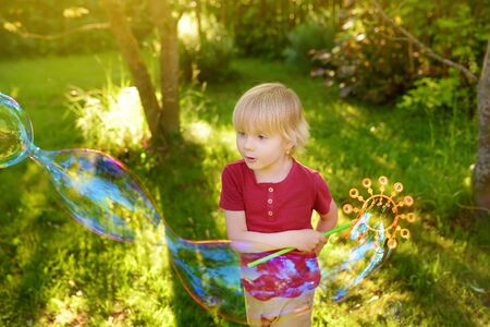 Cute little boy is playing with big bubbles outdoor. Child is blowing big and small bubbles simultaneously. Active summer leisure for kids.の写真素材