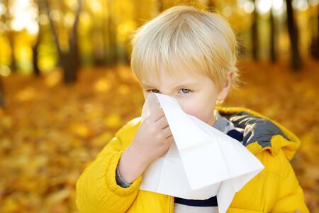 Little boy sneezing and wipes nose with napkin during walking in autumn park. Flu season and cold rhinitis. Allergic kid. Sick kids.の写真素材