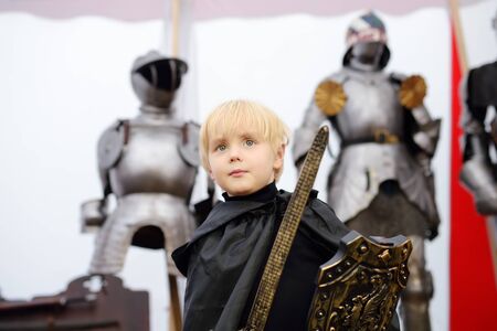 Portrait of a cute little boy dressed as a medieval knight with a sword and a shield on background of knight's armor. Medieval festival or costume party for kidsの写真素材