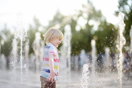 Little boy plays in the square between the water jets in the fountain at sunny summer day. Active summer leisure for kids in the city.の写真素材