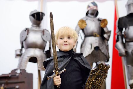 Portrait of a cute little boy dressed as a medieval knight with a sword and a shield on background of knight's armor. Medieval festival or costume party for kidsの写真素材