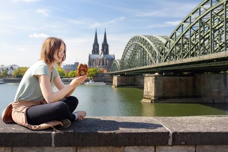 Young woman eats traditional pretzel sitting on embankment of Rhine on background of Cologne Cathedral and Hohenzollern Bridge in Koel, Germany. Tourism and travel by Germany.の写真素材