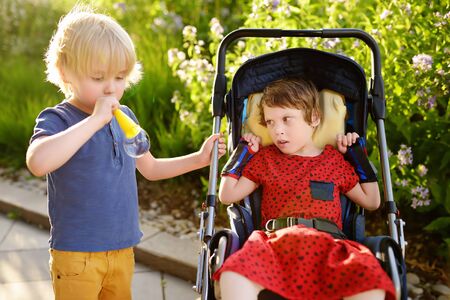 Little boy and disabled girl in a wheelchair playing together in the summer park. Child cerebral palsy. Family with disabled kid.の写真素材