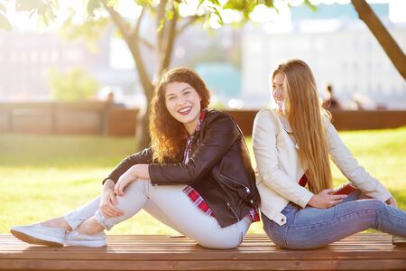 Two beautiful young women talking while sitting on a bench at sunny park. Girlfriends. Communication and gossip.の写真素材