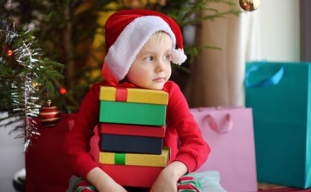 Cute little boy with a Christmas gift under fir tree at home. Portrait of frustrated kid in Christmas morning. Cozy living room with decorated fir treeの写真素材