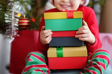 Cute little boy with a Christmas gift under fir tree at home. Portrait of happy kid in Christmas morning. Cozy living room with decorated fir treeの写真素材