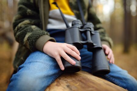 Little boy scout with binoculars during hiking in autumn forest. Child is sitting on large fallen tree and looking through a binoculars. Concepts of adventure, scouting and hiking tourism for kids.の写真素材