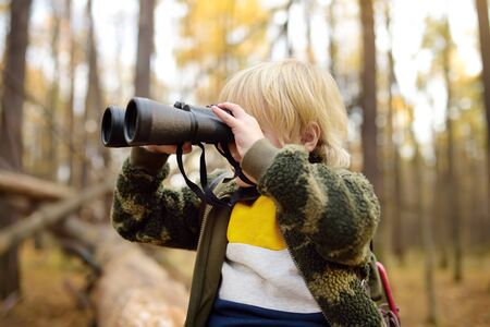 Little boy scout with binoculars during hiking in autumn forest. Child is sitting on large fallen...