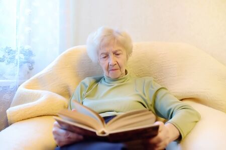 Eldrely woman is reading a book on couch at home. Senior lady lifestyles. Active longevity concepts.