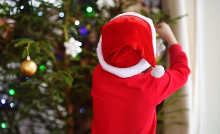 Little boy wearing Santa hat ready for celebrate Christmas. Cute child decorating the Christmas tree with glass toy. Activity for children on winter holidaysの写真素材