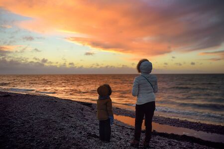 Woman wearing white jacket and little child are walking on Baltic Sea coast on sunset of winter day. Outdoors activity for family with kids in winter. Estonia.の写真素材
