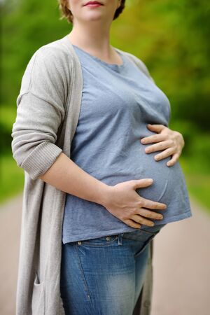 Young pregnant woman during walking in spring park. She supports with hands large pregnant belly.