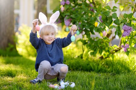 Pretty little boy wearing bunny ears hunting for eggs in spring park on Easter day.Traditional...