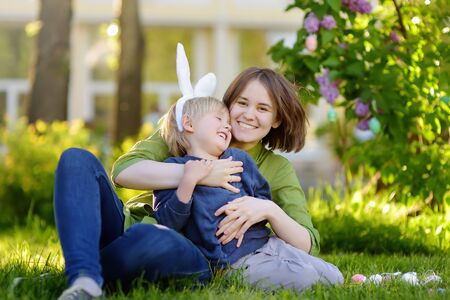 Family celebrate Easter holiday in spring park. Adorable little boy and his charming mother sitting on the grass, embracing and laughing. Traditional easter festival outdoors.の写真素材
