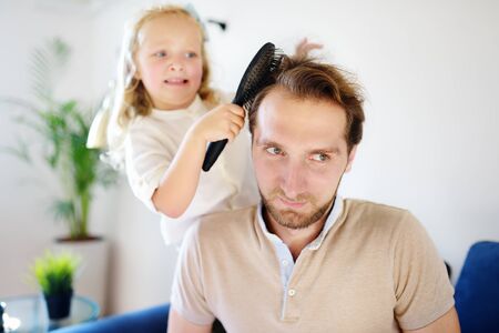 Little curly hair daughter making funny hairstyle for her young handsome father. Preschooler girl...