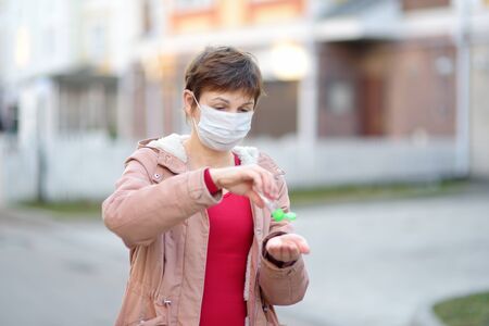 Mature woman wearing medical face mask makes disinfection of hands with sanitizer on city streets or...