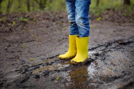 Mischievous preschooler child wearing yellow rubber rain boots jumping in large wet mud puddle. Kid...